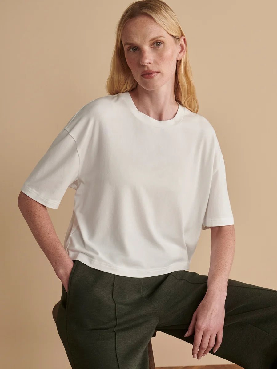 A woman wearing a white short-sleeve crewneck t-shirt and dark olive green trousers, posing against a solid beige background.